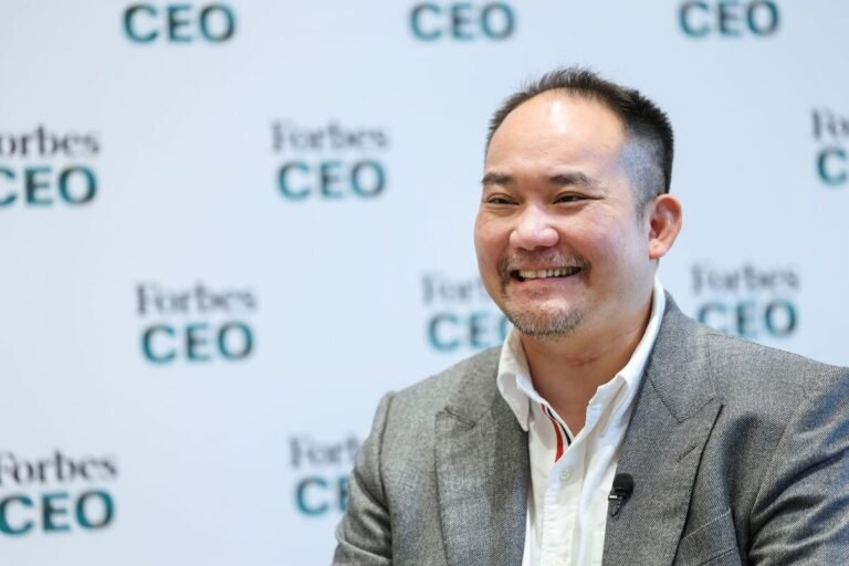 一名男性在印有"Forbes CEO"字样的背景板前微笑，身穿灰色西装外套，胸前佩戴麦克风